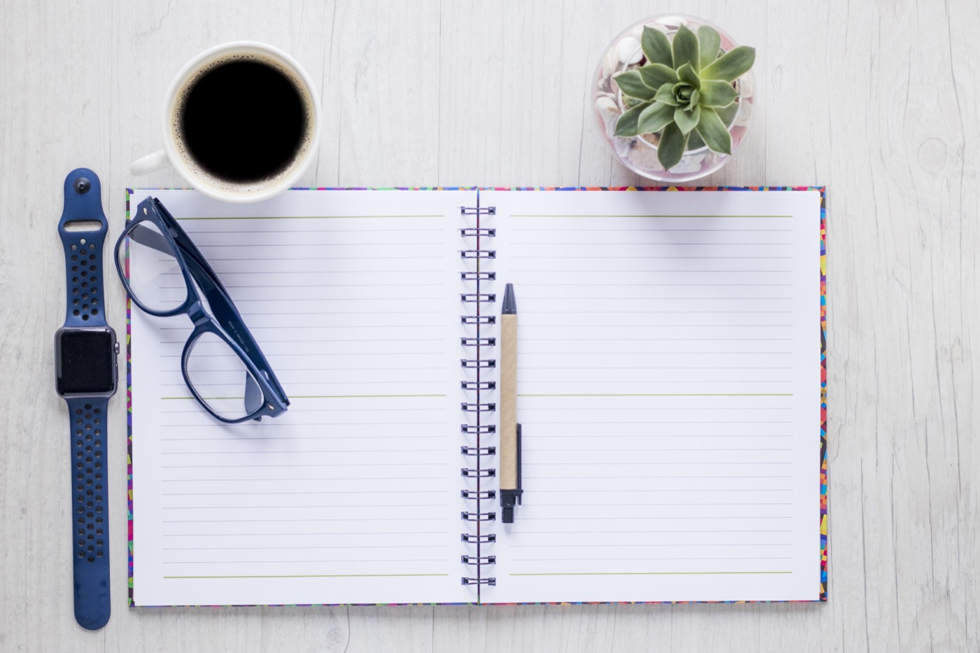 A notebook, coffee, and plant arranged on a desk